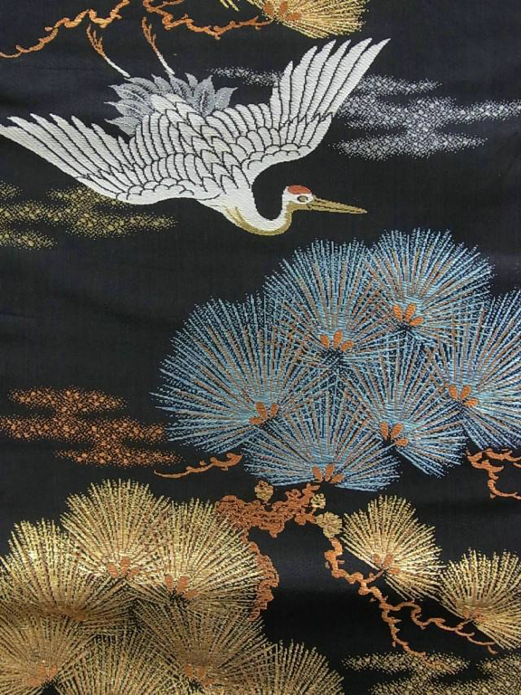 Nagoya obi. Sur un fond de soie noire, une grue en vol au-dessus des pins est tissée selon la technique de tissage Oshima. Le Label « Oshima Ori » est visible quand la ceinture est nouée sur le dos, mais c’est surtout la disposition saisissante de la grue d'un blanc immaculé, des pommes de pin dorées et des aiguilles de pin nuancées de bleu qui nous font apprécier ce tissage délicat.